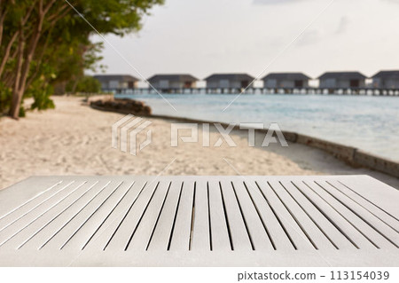 Empty top of white wooden table for product placement and display. Blue lagoon with water bungalows and tropical plants on background. Maldives 113154039