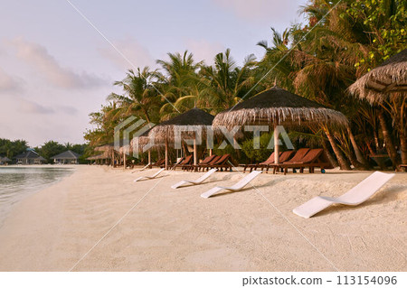 Beach chairs under straw umbrellas. Indian ocean coastline on Maldives island. White sandy beach and calm sea. Travel and vacation concept. 113154096