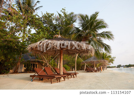 Beach deck chairs under straw umbrellas. Indian ocean coastline on Maldives island. White sandy beach and calm sea. Travel and vacation concept. Beach deck chairs under straw umbrellas. Indian ocean coastline on Maldives island. White sandy beach and calm sea. Travel and vacation concept. 113154107