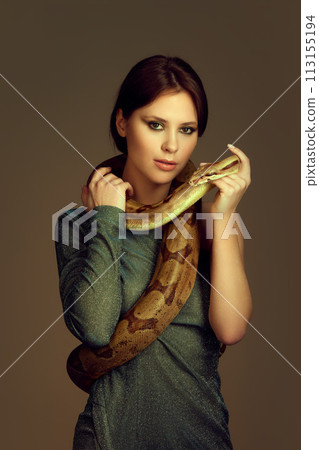 Young woman in glittery green dress with makeup posing with snake against brown studio background. Beauty salon poster 113155194