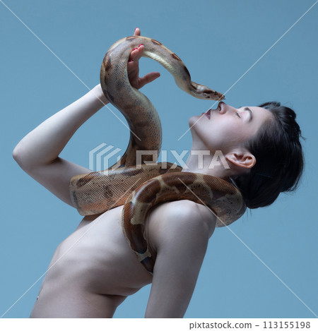 Beautiful brunette young woman with slim body posing with snake close to her face on blue studio background. Kiss of danger 113155198