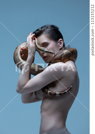 Beautiful brunette young woman with slim body posing with snake on blue studio background. Inner self and freedom Beautiful brunette young woman with slim body posing with snake on blue studio background. Inner self and freedom 113155212