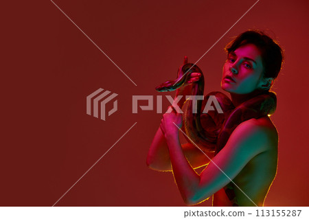 Portrait of sensual, beautiful young woman in slim body in underwear standing with snake against red background in neon light. Passion Portrait of sensual, beautiful young woman in slim body in underwear standing with snake against red background in neon light. Passion 113155287