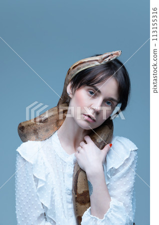 Portrait of elegant young woman in white dress, snake draped over shoulders, intense gaze, posing against blue-gray background. 113155316