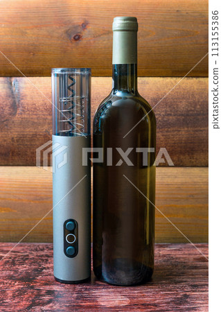 An electric corkscrew, a bottle of wine and a bottle opener lie on a dark wood background 113155386