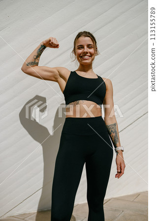 Smiling woman athlete raising arm and showing biceps while standing on modern buildings background Smiling woman athlete raising arm and showing biceps while standing on modern buildings background 113155789