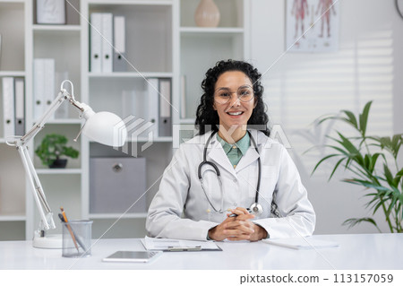 Professional Hispanic female doctor sitting at her desk with a stethoscope, ready to start a video call in a clinic office setting. 113157059