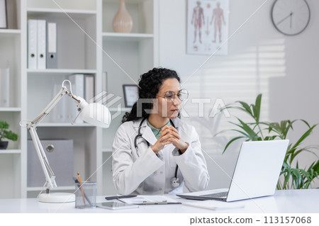 Thoughtful Hispanic female doctor sitting at her desk in a bright medical office, looking at a laptop screen. 113157068