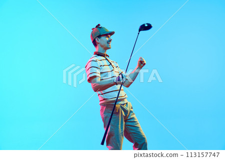 Overjoyed golf player in retro clothes and cap raising hands after perfect goal in neon light against gradient blue background. 113157747