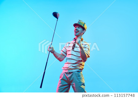 Joyful golfer in retro outfit and cap celebrating perfect goal with raised fist and golf club in neon light against gradient blue background. 113157748