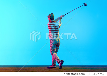 Back view portrait of young man, professional golfer playing golf on green lash grass in neon light against gradient blue background. Back view portrait of young man, professional golfer playing golf on green lash grass in neon light against gradient blue background. 113157752