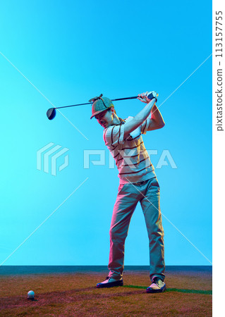 Young athletic man, professional golfer playing golf on green lash grass in neon light against gradient blue background. 113157755