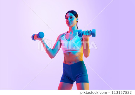 Young athlete woman doing exercises with dumbbells for biceps in neon light against gradient studio background. 113157822