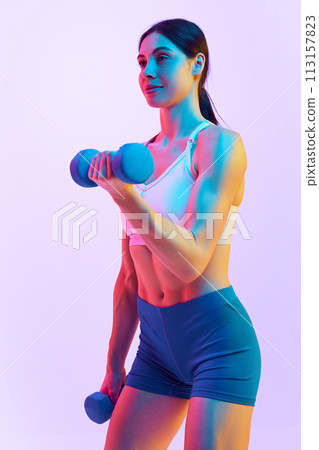 Young attractive, fit woman doing exercises with dumbbells for biceps in neon light against gradient studio background. Young attractive, fit woman doing exercises with dumbbells for biceps in neon light against gradient studio background. 113157823