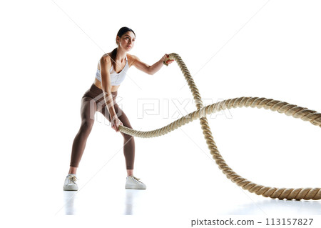 Young strong fit woman doing exercises with battle ropes at gym against white studio background. 113157827
