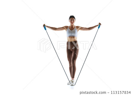 Female athlete in sportswear using resistance band for training against white studio background. 113157834