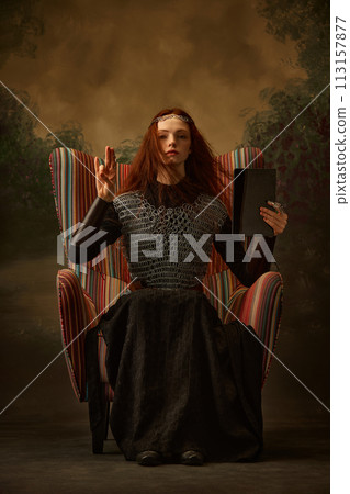 Young woman, dressed as brave knight sitting on chair and holds modern tablet with blank screen against vintage studio background. Young woman, dressed as brave knight sitting on chair and holds modern tablet with blank screen against vintage studio background. 113157877