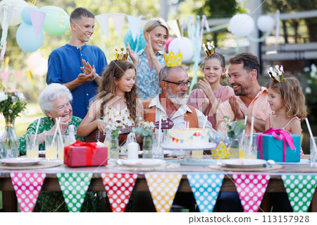 Garden birthday party for elderly man. Handsome senior birthday man getting cake gift from family. Garden birthday party for elderly man. Handsome senior birthday man getting cake gift from family. 113157928