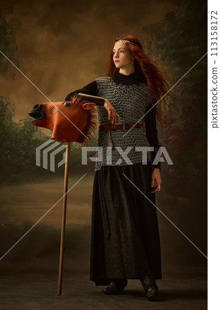 Red-haired princess in chainmail and black dress touching whimsical horse head on stick, toy against vintage studio background. Red-haired princess in chainmail and black dress touching whimsical horse head on stick, toy against vintage studio background. 113158172