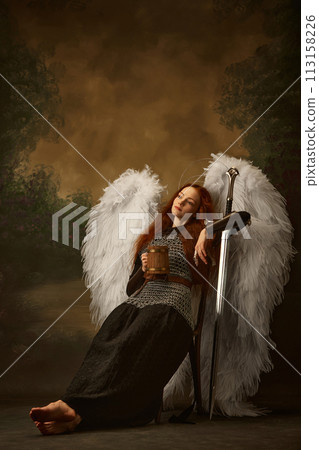Redhead woman looks as angel in chainmail with sword, with large white wings and golden halo drinking beer against vintage studio background. Redhead woman looks as angel in chainmail with sword, with large white wings and golden halo drinking beer against vintage studio background. 113158226