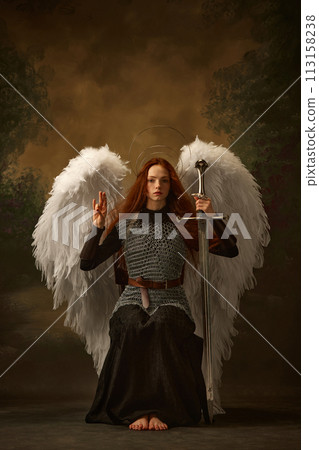 Redhead angel in chainmail, raising sword, with large white wings and golden halo against vintage studio background. Renaissance-style Redhead angel in chainmail, raising sword, with large white wings and golden halo against vintage studio background. Renaissance-style 113158238