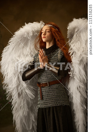Red-haired maiden in angelic wings and medieval attire embodies a serene, prayerful stance against vintage studio background. Red-haired maiden in angelic wings and medieval attire embodies a serene, prayerful stance against vintage studio background. 113158285