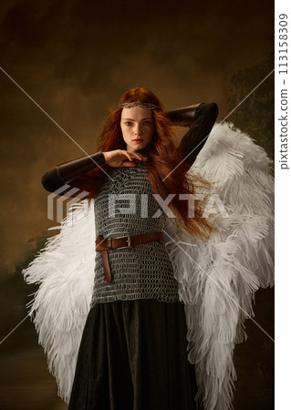 Red-haired woman with angel wings and chainmail, wearing a diadem, poses dramatically against vintage studio background. Red-haired woman with angel wings and chainmail, wearing a diadem, poses dramatically against vintage studio background. 113158309
