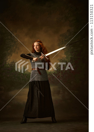 Full-length portrait of young princess in chainmail with sword posing showing her brave against vintage background. Full-length portrait of young princess in chainmail with sword posing showing her brave against vintage background. 113158321