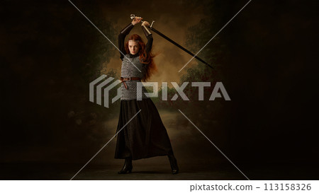 Portrait of beautiful young woman dressed as medieval person, knight and posing raising sword against vintage background. 113158326