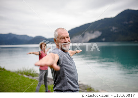 Senior couple doing outdoor yoga, tai chi, pilates by the lake in the autumn. Elderly husband and wife spending active vacation in the mountains. Pensioners stretching after outdoor workout Senior couple doing outdoor yoga, tai chi, pilates by the lake in the autumn. Elderly husband and wife spending active vacation in the mountains. Pensioners stretching after outdoor workout 113160017