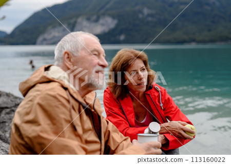 Active elderly couple hiking together in mountains. Drinking coffee and having healthy snack, to get energy. Senior tourist with backpacks resting during hike. 113160022