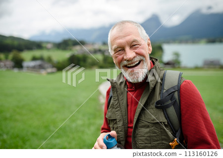 Portrait of smiling elderly man walking outdoors with trekking poles, going on hiking trail. Senior tourists enjoying view Portrait of smiling elderly man walking outdoors with trekking poles, going on hiking trail. Senior tourists enjoying view 113160126