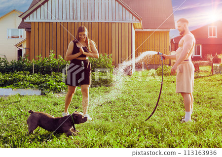 Playful dog grabs splashes of water from garden hose with its mouth during an evening walk in courtyard of country house with young European man and woman. Playful dog grabs splashes of water from garden hose with its mouth during an evening walk in courtyard of country house with young European man and woman. 113163936