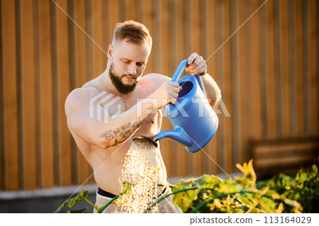 An athletic man waters vegetable beds using garden watering can. 113164029