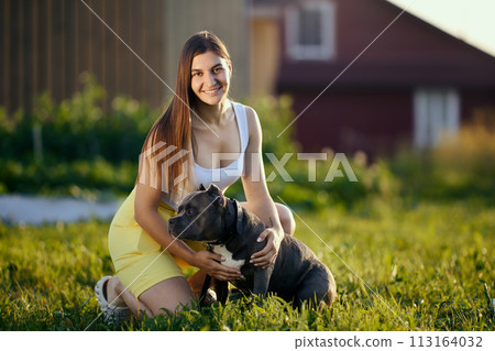 Exterior portrait of young European woman with her American Bully breed dog on summer evening in village. 113164032