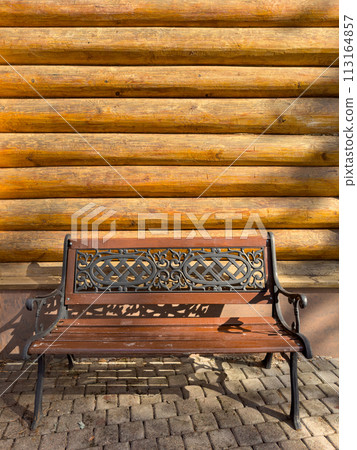 A wrought-iron bench stands next to a log building, Logs of a wooden house in close-up on a sunny day, a bathhouse, the hut A wrought-iron bench stands next to a log building, Logs of a wooden house in close-up on a sunny day, a bathhouse, the hut 113164857