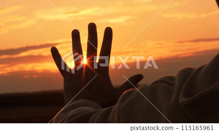 Silhouette of hand at orange sunset. Man stretches hand towards sun, warming palm in summer rays. Unity with nature, enjoyment of trip. Guy gains strength, contemplating, enjoying solitude with nature 113165961