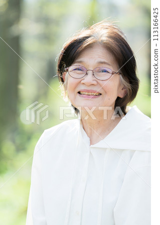 Close-up of a natural smiling senior woman looking at the camera outdoors in backlit fresh greenery Close-up of a natural smiling senior woman looking at the camera outdoors in backlit fresh greenery 113166425