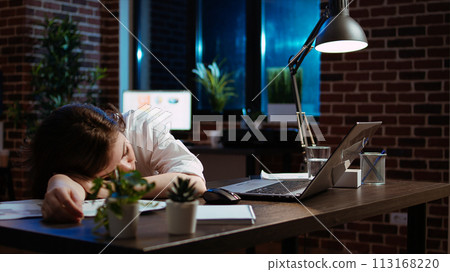 Sleepy employee asleep on computer desk late at night in brick wall office. Panning shot of tired businesswoman sleeping with head on table while working overnight on laptop, camera A 113168220