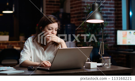 Weary accountant struggling to remain awake at workspace desk while inputting data on laptop, panning shot. Close up of exhausted businesswoman yawning in office while working overnight, camera B 113168221