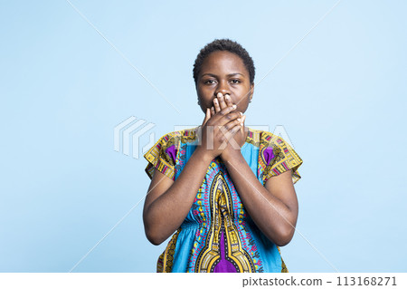 African american model covering her mouth to be silent, recreating three wise monkeys symbol to keep quiet in front of the camera. Woman showing inspirational traditional sign in studio. 113168271