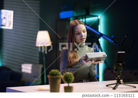 Young entertainer in neon lit studio doing technology review of bluetooth portable speaker for online streaming channel. Influencer films music playing device unboxing for generation Z audience Young entertainer in neon lit studio doing technology review of bluetooth portable speaker for online streaming channel. Influencer films music playing device unboxing for generation Z audience 113168321