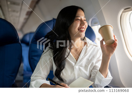 An elegant Asian businesswoman is sipping coffee while looking at the view outside the plane window. An elegant Asian businesswoman is sipping coffee while looking at the view outside the plane window. 113169068