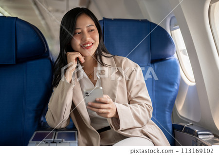 An attractive Asian businesswoman is sitting at the window seat with a smartphone in her hand. An attractive Asian businesswoman is sitting at the window seat with a smartphone in her hand. 113169102