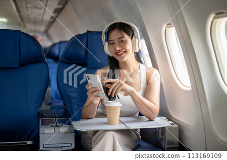 An attractive woman is using her phone and listening to music on her headphones during the flight. 113169109