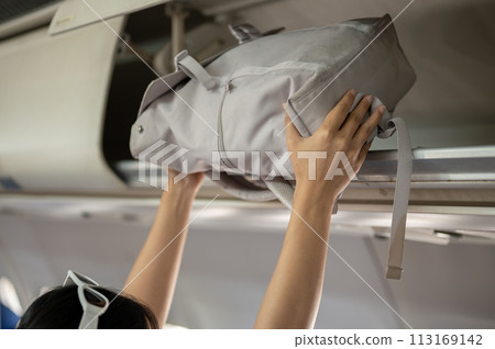 A close-up image of a female passenger is putting her bag in an overhead bin, traveling by plane. A close-up image of a female passenger is putting her bag in an overhead bin, traveling by plane. 113169142