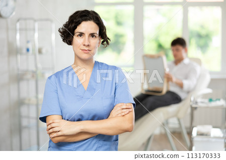 Middle-aged woman doctor posing in doctor's cabinet with patient lying on clinical chair 113170333