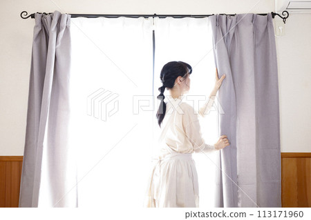 A woman in an apron standing by the window and opening the curtains 113171960