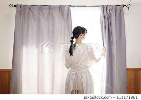 一個穿著圍裙的女人站在窗邊打開窗簾 113171961