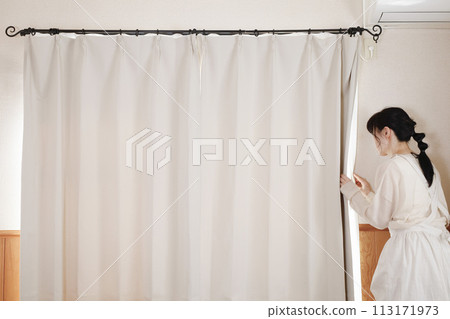 一個穿著圍裙的女人透過窗簾之間的縫隙向外看 一個穿著圍裙的女人透過窗簾之間的縫隙向外看 113171973